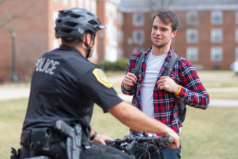 Male student in plaid shirt and wearing backpack talks to police officer on motor scooter 